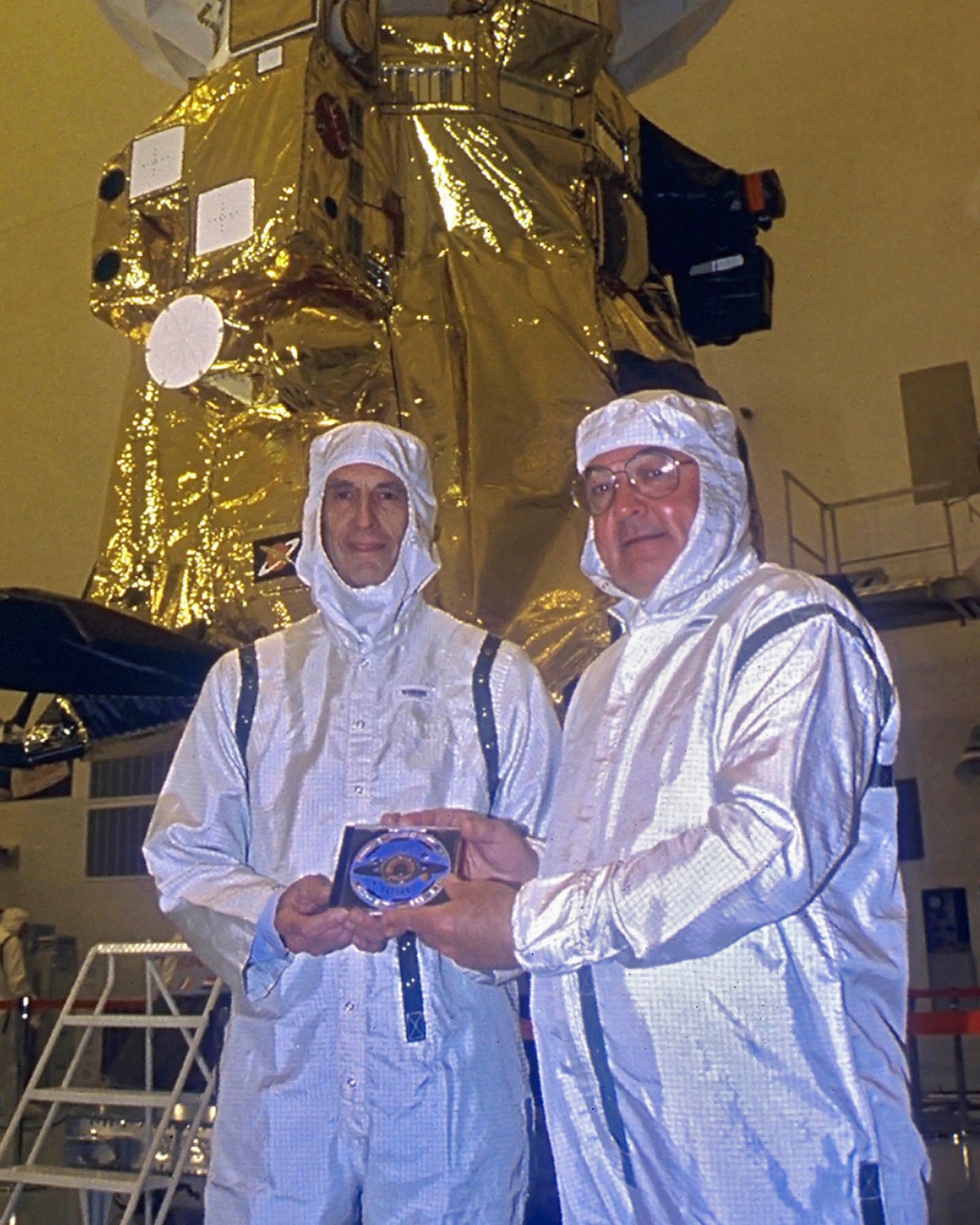 In the Payload Hazardous Servicing Facility at the Kennedy Space Centre Kohlhase (left) and Cassini Programme Manager, Richard Spehalski, hold the DVD containing 616,400 digitized signatures of people from 81 countries which was sent to Saturn on board Cassini, seen behind them. Charley designed the disk with the flags of 28 nations and symbolic golden eagle wing feathers. Credit: NASA/JPL